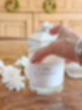 Hand placing a Lime + Gardenia candle on a rustic wooden table, with white flowers and a white ceramic jar in the background.
