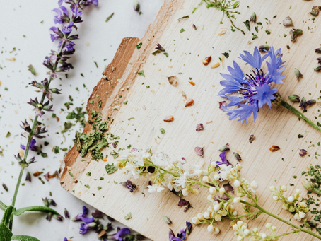 Dried purple and white flowers scattered on a wooden board, with green herbs and seeds, creating a rustic, natural scene.