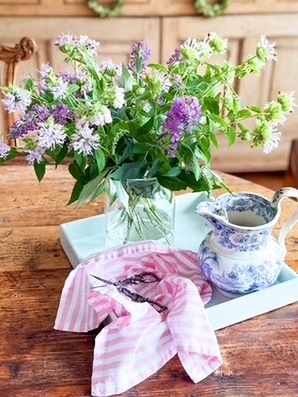 Purple wildflowers in a glass vase on a wooden table, beside a floral teapot and pink-striped cloth on a blue tray.