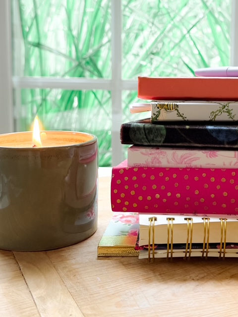 Lit candle beside a stack of colorful notebooks on a wooden table by a bright window with green foliage.