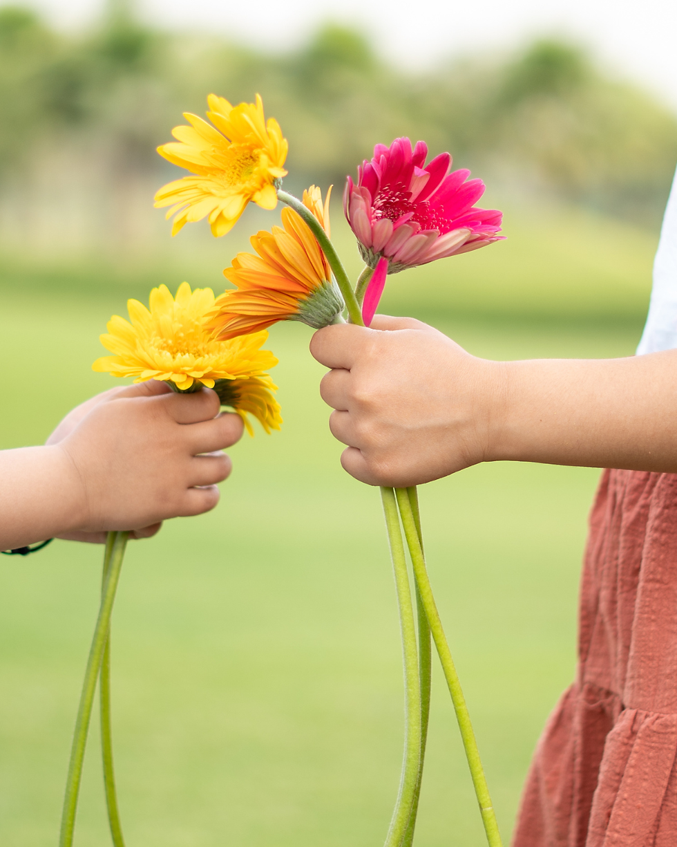 Two children’s hands hold bright yellow, orange, and pink gerbera daisies against a soft green outdoor background.