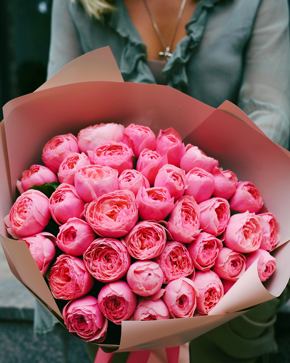 A person holds a bouquet of vibrant pink peonies wrapped in light pink paper. The soft green blouse adds to the warm, cheerful mood.