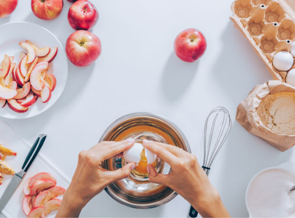 Hands cracking an egg into a bowl on a white table. Surrounding are apples, sliced apples, a whisk, flour, and an egg carton. Bright setting.
