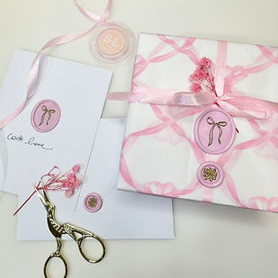 a flat lay of a gift wrapped present covered in a print of bows with two wax seals and a card with a bow wax seal on it and some baby's breath with scissors