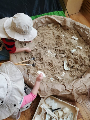 Two PreK children wearing safari hats and role playing as paleontologists as they dig in a round circle of sand in a classroom.