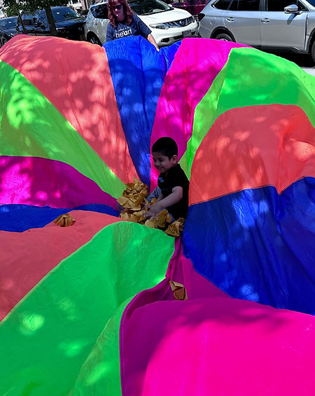 A pre-k boy in the middle of a giant multi colored sheet on the floor.