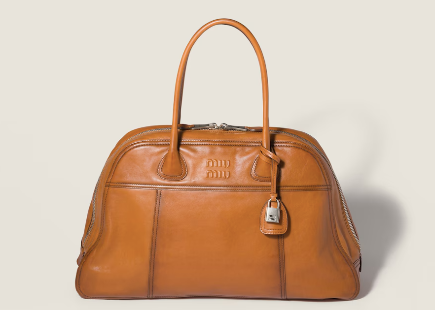 Bolsa de couro castanha claro com o logotipo Miu Miu, zíper duplo e alça superior. Fundo bege claro, design elegante e sofisticado.