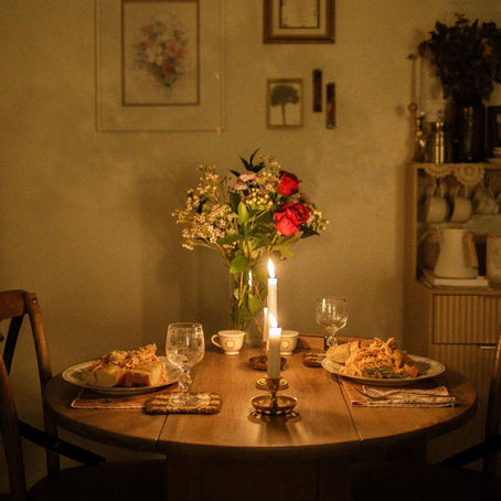 Mesa de jantar romântica à luz de velas, com duas refeições servidas em pratos, copos de vinho, flores com rosas vermelhas ao centro e decoração acolhedora, criando um ambiente íntimo e aconchegante para um jantar a dois.