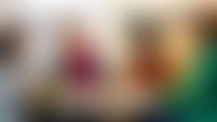 Four women sit at a table in an office, chatting and laughing over coffee. One wears a pink scarf; another, a green blazer. Mood is cheerful.