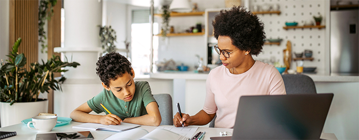 Woman working on laptop while son does schoolwork nearby.