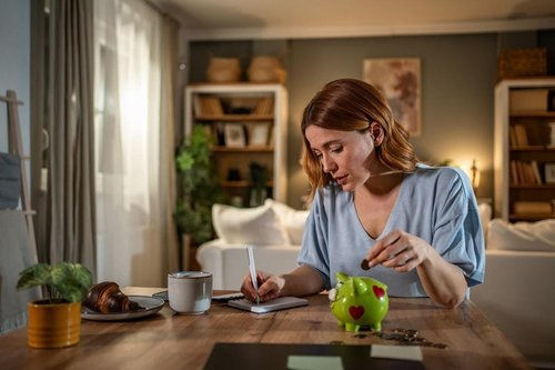 Woman putting coin in piggy bank while writing in notebook