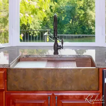 Copper apron front workstation sink with warm swirled patina under a large window