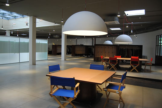Show-room a forma circolare caratterizzato da pozzo di luce con lucernari, tavoli da esposizione in legno, mobili da esposizione in ferro corten. Illuminazione puntiforme e lampade a sospensione costituite da una cupola semisferica bianca. Pavimentazione in cemento spazzolato.