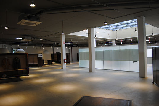 Show-room a forma circolare caratterizzato da pozzo di luce con lucernari, tavoli da esposizione in legno, mobili da esposizione in ferro corten. Illuminazione puntiforme e lampade a sospensione costituite da una cupola semisferica bianca. Pavimentazione in cemento spazzolato.