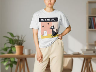 Woman with shaved head in a white ME & MY BOO T-shirt with a black cat and ghost graphic, standing in a bright home office.