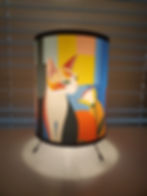 Colorful lamp with abstract cat and flower design. Background has blinds. Soft white light on a white surface creates a calm mood.