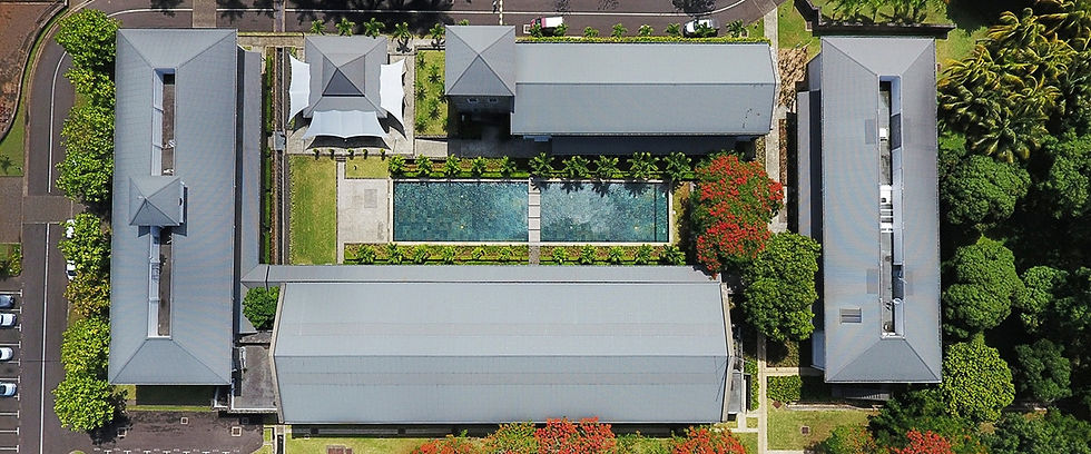 Aerial view of a complex with four gray-roofed buildings surrounding a rectangular pool, lush greenery, and red-flowering trees.