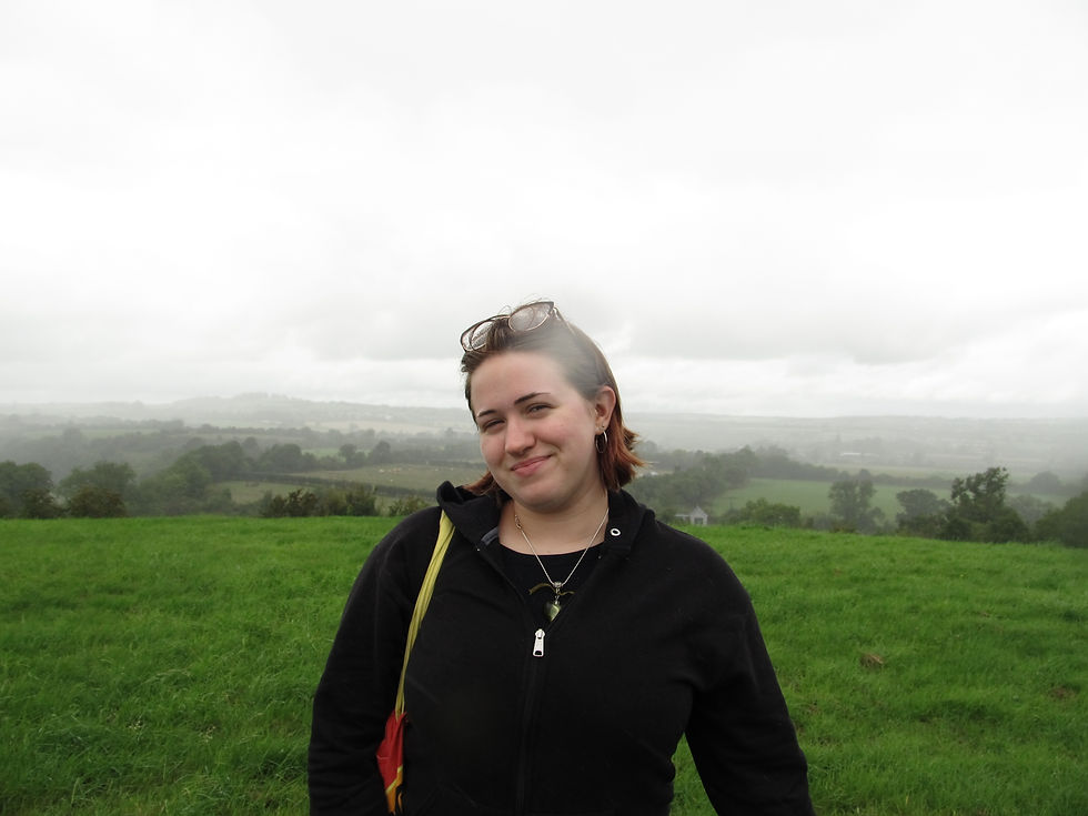 Smiling woman in a black jacket stands on a grassy hill with misty green countryside and cloudy sky behind her.