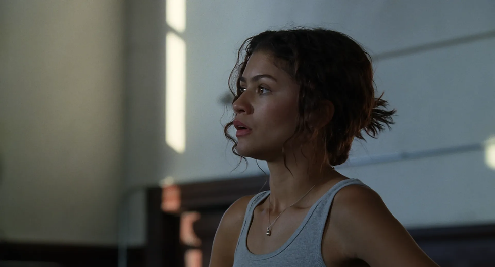 Woman (Zendaya) in a gray tank top looks left in a dim room with sunlight streaks, appearing tense and focused.
