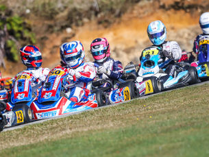 Go-kart racers in colorful helmets and suits compete on a track. Numbers 50, 25, and 21 visible on karts. Energetic and competitive scene.