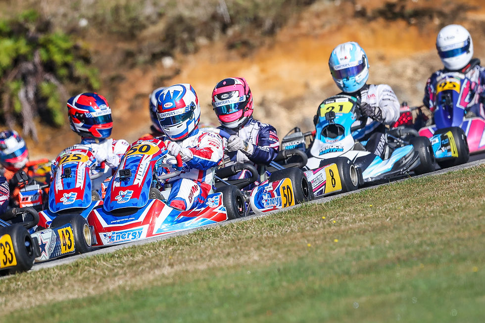Go-kart racers in colorful helmets and suits compete on a track. Numbers 50, 25, and 21 visible on karts. Energetic and competitive scene.