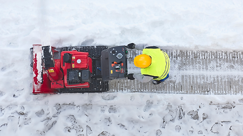 Worker in a yellow hard hat and vest using a red snow blower to clear a snowy sidewalk.