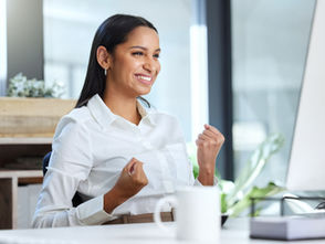 Smiling woman in a white shirt clenches fists in excitement at a desk. Bright office setting with plants and a white mug nearby.