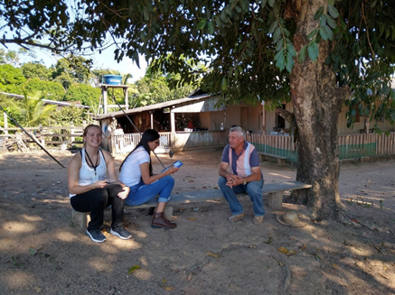 Rachel, a student interviewer, and a farmer sitting on a low bench under a tree.