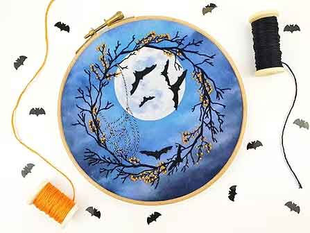 Halloween embroidery design of bats flying across a full moon with a wreath of branches and webs on a blue background, surrounded by supplies.