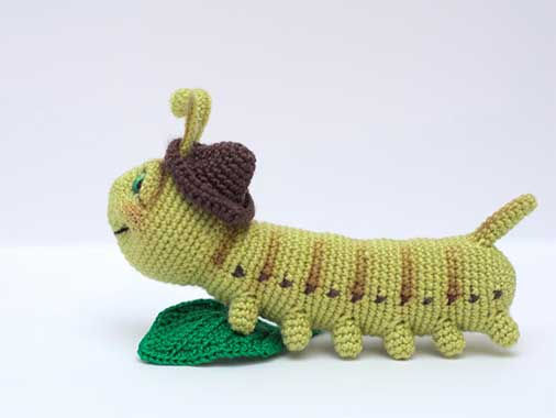 Small green amigurumi caterpillar with brown head cap and multiple legs standing on crocheted leaf.