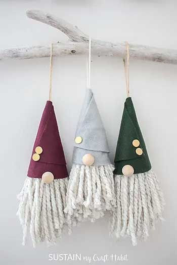 DIY gnome ornaments with yarn beards and felt hats in burgundy, gray, and green, hanging on a rustic branch.