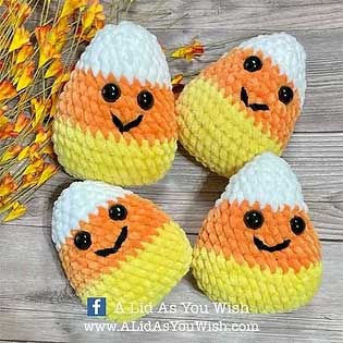 Four smiling plush candy corn made from a candy corn crochet pattern, displayed on a wooden surface with fall accents.