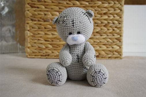 Crochet teddy bear pattern in light gray with stitched details, blue nose, and soft body, sitting on a neutral surface with a wicker basket in the background.
