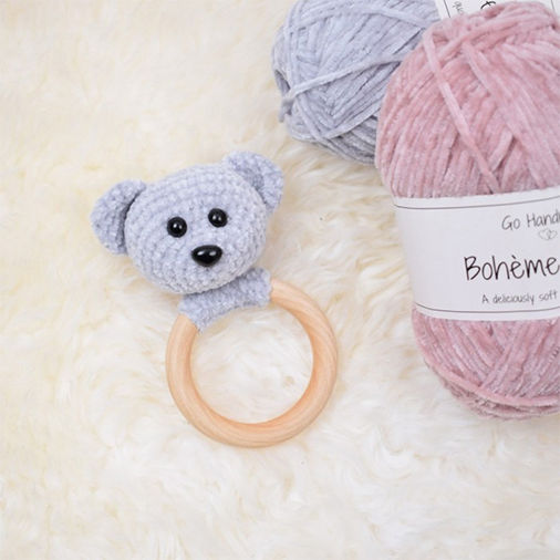 Crochet pattern teddy bear rattle with a gray bear head on a wooden ring, displayed alongside pink and gray yarn skeins.
