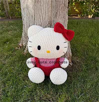 Hello Kitty crochet plush sitting outdoors by a tree, wearing a red bow and overalls.