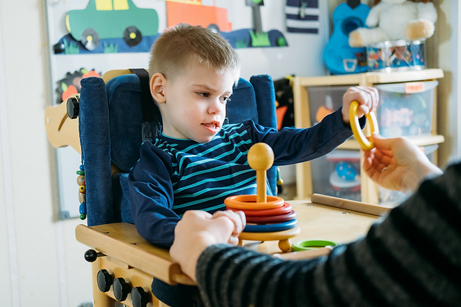 A young child sitting in an activity chair with a tray. They are placing a ring on a stacking toy with an adult hand holding theirs.