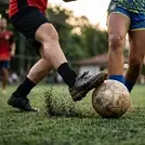 Close-up dinâmico dos pés de dois jogadores em uma disputa de bola agressiva no futebol Society. Um jogador, vestindo bermuda e meias pretas, chuta com força uma bola de couro surrada e suja no gramado sintético verde. O impacto levanta uma nuvem visível de borrachinhas pretas do gramado. À direita, outro jogador, com uniforme de estampa azul e amarela, tenta interceptar, com o pé direito posicionado atrás da bola. O fundo é um parque gramado com árvores e a luz dourada do fim da tarde, criando um efeito de desfoque suave (bokeh).