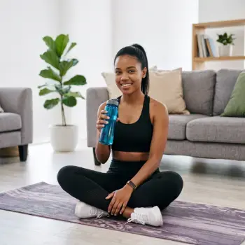Mulher jovem de cabelo preto amarrado em um rabo de cavalo, vestindo roupa fitness preta, sentada de pernas cruzadas sobre um tapete de treino na sala de casa. Ela está sorrindo, olhando para a câmera e segurando uma garrafa de água azul.