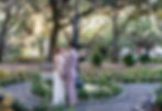 A couple in formal attire holds hands, smiling by a fountain in a sunlit park. Tall trees and benches are in the background.