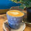 Thumbnail: A denim blue coffee cup filled with a latte, adorned with a leaf design, and a biscuit resting on a white saucer. Beside it is a green, leafy plant.