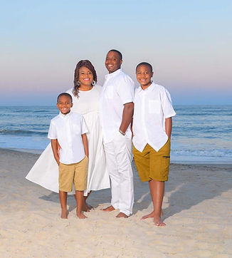 Family of 4 posing for portrait on the beach with a colorful sky in the background.