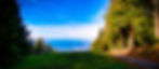 Vue panoramique d'un champ verdoyant bordé d'arbres, lac lointain et ciel bleu.