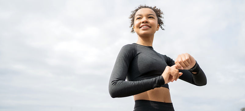 Une jeune femme souriante vérifie sa montre, en plein air, journée nuageuse.