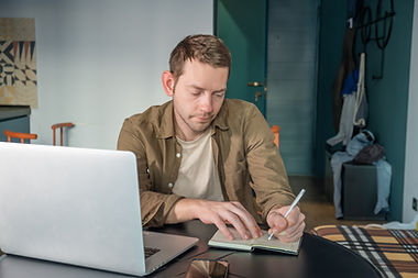 man-writing-notes-notebook-while-working-laptop-home-office-table-male-digital-nomad.jpg