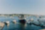 Boats docked at a marina, with a large ferry center. Blue sky, calm water, and trees in the background. A tranquil, summery scene.