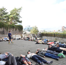 a group of people lie down stretching and forming a wide circle with their bodies side by side. João stands to guide the experience.