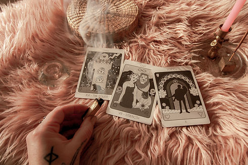 Three tarot cards sit on a pink fluffy blanket, a hand holds a smoking piece of palo santo over top of them