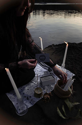 A woman sits on a rock by the ocean flipping tarot cards onto a black and white cloth with lit candles around her