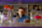 Caucasian man in a blue plaid shirt looking toward the camera through a shelf of labeled glass bottles