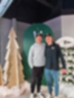Two men smiling at a holiday event with wooden tree decor.